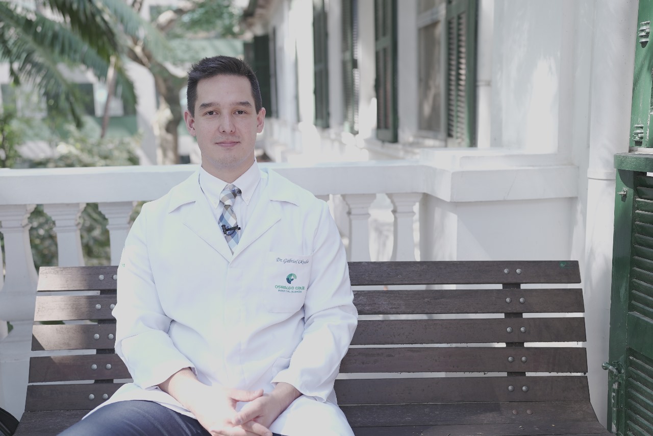 Gabriel Okuda é um homem branco que está de jaleco sentado em um banco de madeira